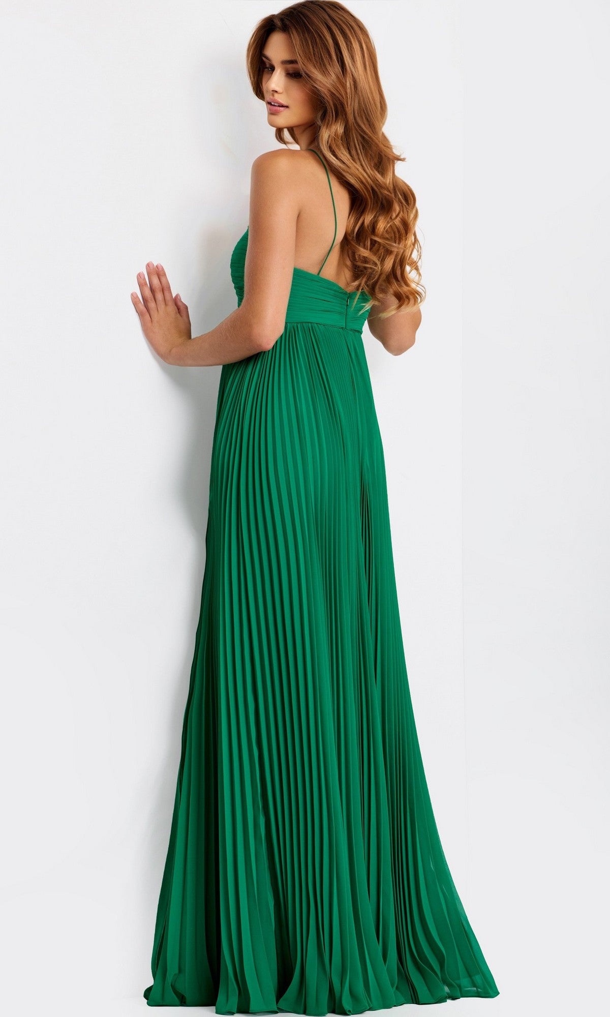 A woman with long, wavy hair stands facing a white wall and looks over her shoulder, wearing the floor-length green Jovani 47820 prom dress with pleats.